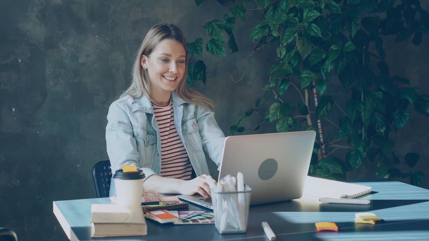 Woman smiles while working on laptop at desk with plant.