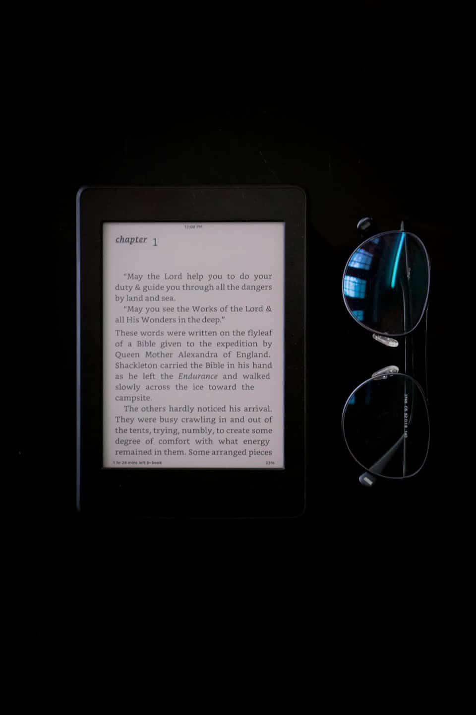 An e-reader and round glasses rest on a dark surface.